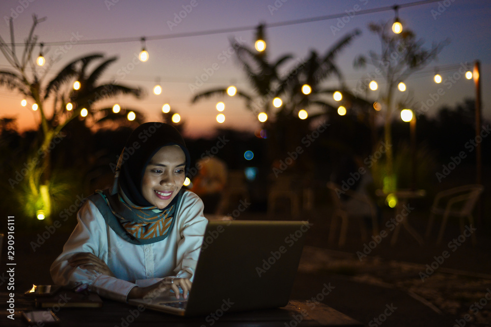 Smiling business asian muslim woman browsing on laptop at outdoor cafe in evening.