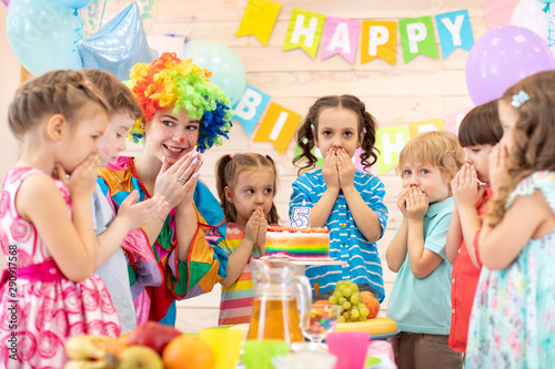 Children with clown celebrating birthday party. Group of kids stand arround festive table waiting surprise