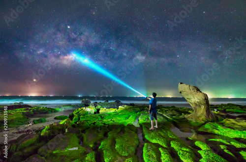 Night landscape with milky way and silhouette of a happy man standing beside mossy rocks on the beach
