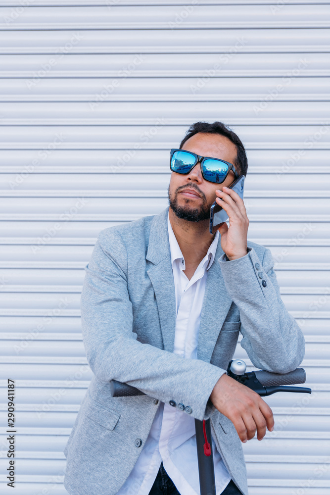 Latin adult man with sunglasses, well dressed and electric scooter talking on his mobile phone sitting on the street with a white wall background Latin adult man with sunglasses, well dressed and electric scooter talking on his mobile phone sitting on the street with a white wall background