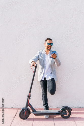 Latin adult man with sunglasses, well dressed and electric scooter talking on his mobile phone sitting on the street with a white wall background