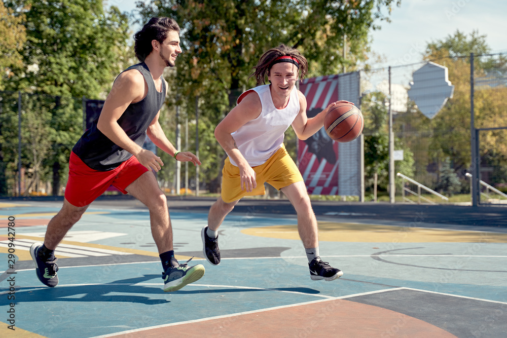 Photo of two sporty men playing basketball on playground on summer day.