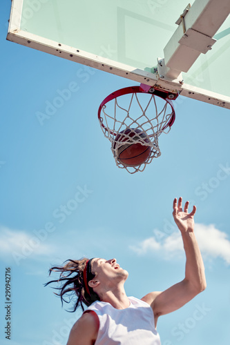 Photo of sports man throwing ball into basketball hoop on sports field on street on summer day.