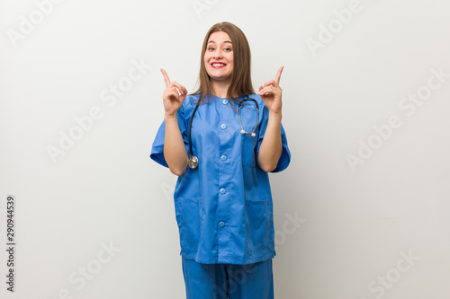 Photos Young nurse woman against a white wall indicates with both fore fingers up showing a blank space