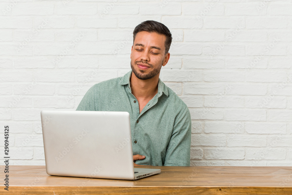Young filipino man sitting working with his laptop touches tummy, smiles gently, eating and satisfaction concept. Young filipino man sitting working with his laptop touches tummy, smiles gently, eating and satisfaction concept.