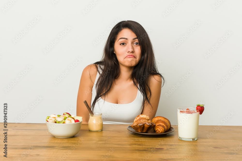 Young curvy woman taking a breakfast shrugs shoulders and open eyes confused. Young curvy woman taking a breakfast shrugs shoulders and open eyes confused.