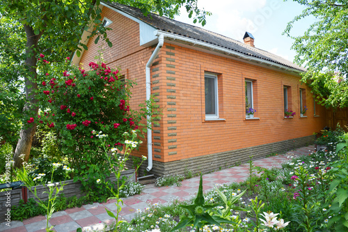 Beautiful brick house with asbestos roof, rain gutter, pathway in cozy garden with trees and roses