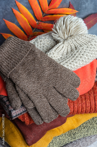 Stack of folded warm knitted women's sweaters, scarf, hat with pompom, gloves in warm colors and bright autumn leaves. Closeup