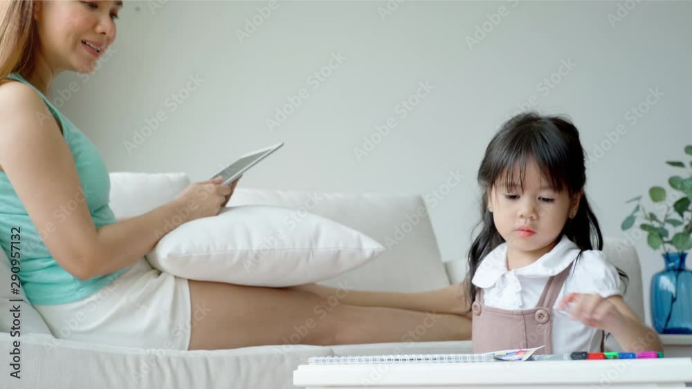 An Asian mother playing with a tablet totally concentrating with the social network and ignoring her daughter practicing drawing by herself An Asian mother playing with a tablet totally concentrating with the social network and ignoring her daughter practicing drawing by herself