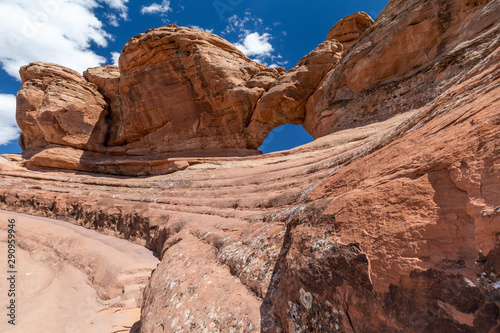 Wide angle view of an imposing red sandstone rock formation with a natural arch on top, under a blue sky with puffy clouds © Roberto