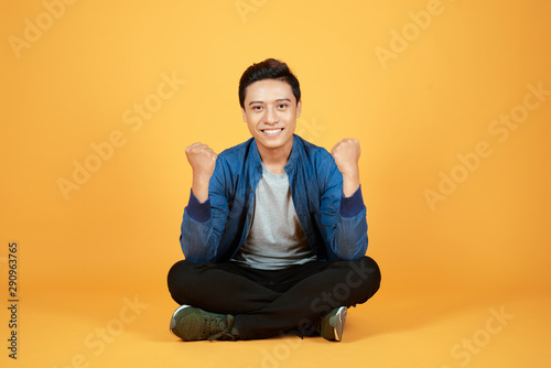 Handsome cheerful young Asian guy wearing casual jeans denim, shoes, sitting on floor with crossed legs, raising hands up. Isolated on orange background © makistock