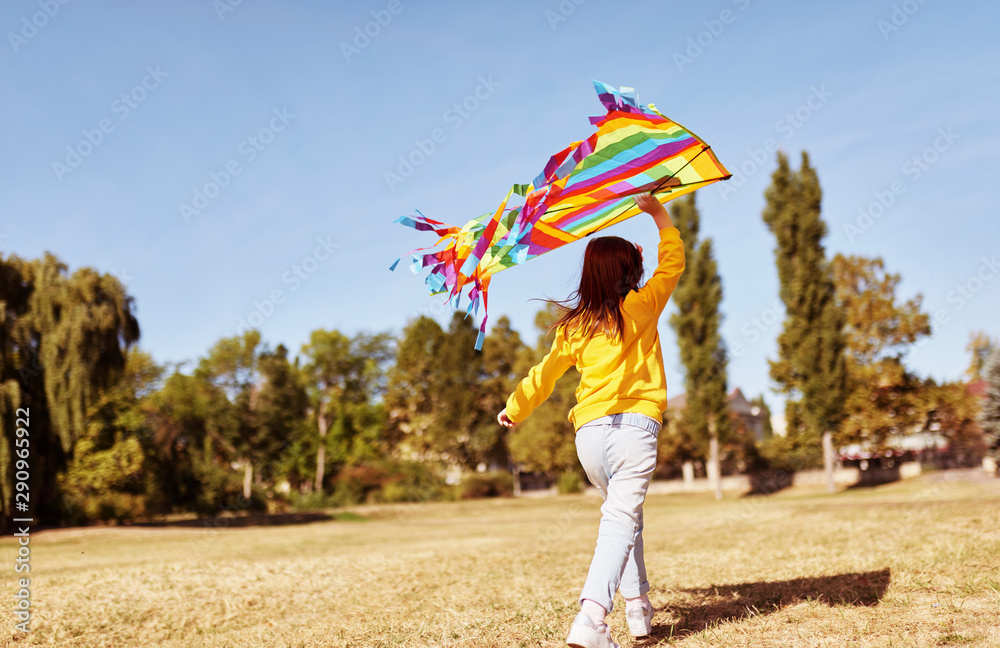 Rear view horizontal image of happy child little girl wearing yellow sweater, running with a big colorful kite on a sunny day outdoors. Cute kid playing and have fun outside with a kite in the park.