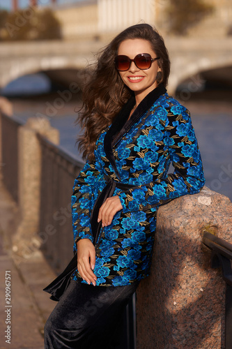 Fashion style outdoor portrait of young beautiful caucasian woman with long wavy brunette hair in blue colorful coat and black trousers. Female model in suit photo