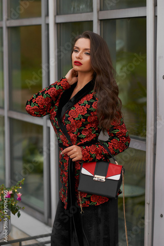 Body length outdoor portrait of young beautiful caucasian woman with long wavy brunette hair in red coat, black trousers and sunglasses with leather handbag posing againsy modern glass and metal wall photo