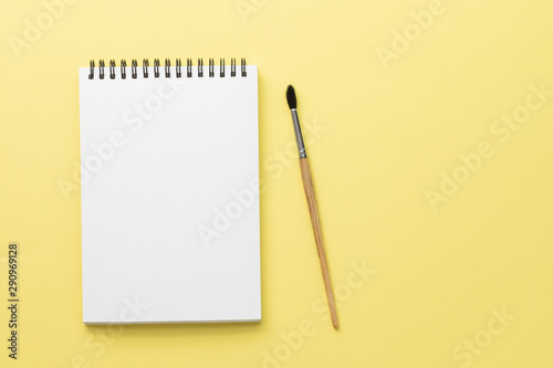 White clean notebook on black spring with wooden squirrel brush on bright yellow paper background with copy space, top view. Artistic workspace