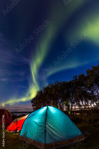Night shot of an icelandic landscape with colorful camping tents in the foreground and silohuette of trees in the background, under a blue sky with green northern lights