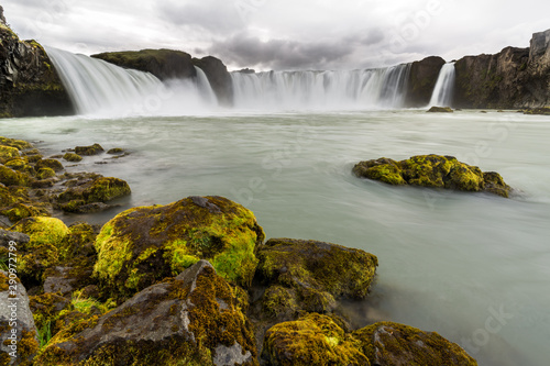 Icelandic landscape with moss-covered rocks in the foreground and Godafoss  one of the great waterfalls  in the background  under a cloudy sky