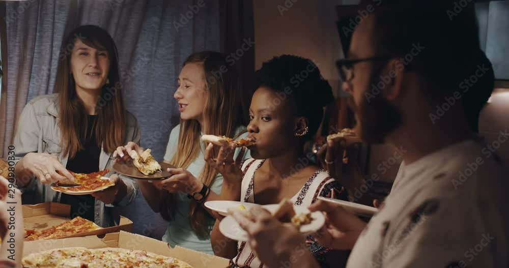 Beautiful young multiethnic women chat, smile eating pizza hanging out with friends at kitchen house party slow motion.