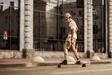 Happy man in white shirt ride on skateboard near road on street background. Outdoors portrait of stylish guy. Urban lifestyle