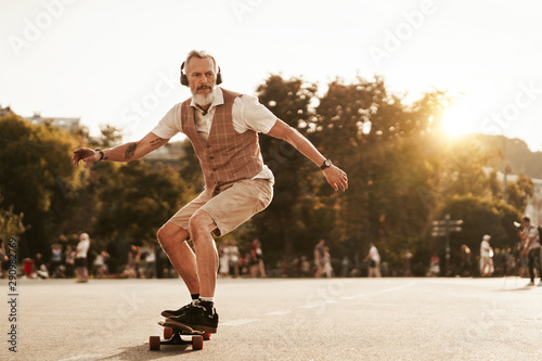 Skater posing in sunset evening on city street. Stylish hipster man with longboard ride near road on buildings background. Portrait of sport male model in urban style. Traveler. Tourist