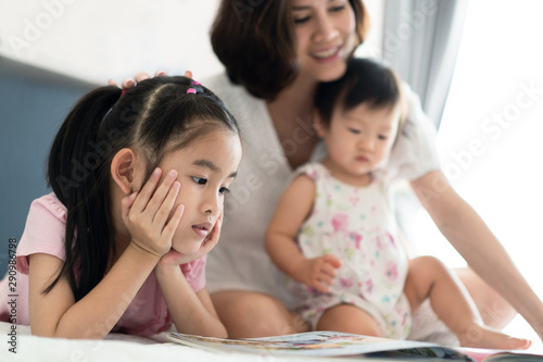 Beautiful Asian mother reading fairly tale story book with her children on the bed. Mom pointing the book telling story while their kids pay attention on listening. Big sister feeling enjoy and smile.