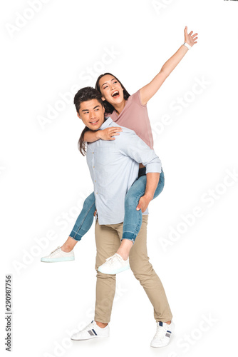 cheerful asian woman waving hand while piggybacking on happy boyfriend on white background