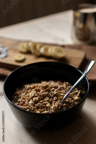 Bowl of granola with saucer of yogurt, banana and coconut chips on light brown wooden board for healthy breakfast. Top view