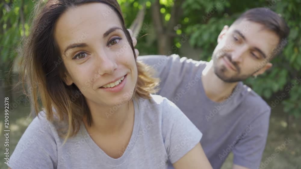 Portrait of young man and woman relaxing in summer garden. The guy touching hair of his wife, she smiling looking at camera. Happy young family resting outdoors. Concept of relationship, tenderness Portrait of young man and woman relaxing in summer garden. The guy touching hair of his wife, she smiling looking at camera. Happy young family resting outdoors. Concept of relationship, tenderness