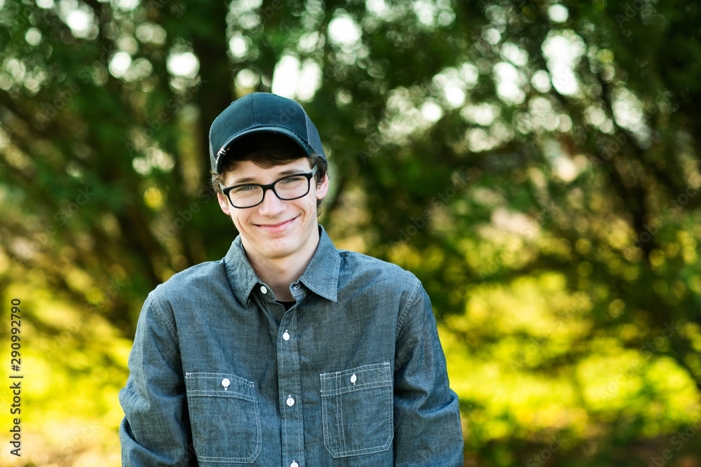Teenage boy with glasses standing outside in front of a tree wearing a gray button up long sleeved shirt and denim jeans with baseball cap on