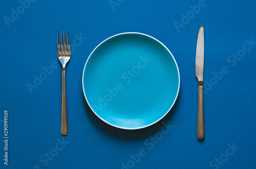 Ready to order. High angle shot of empty color plate, fork, knife lying against blue background.