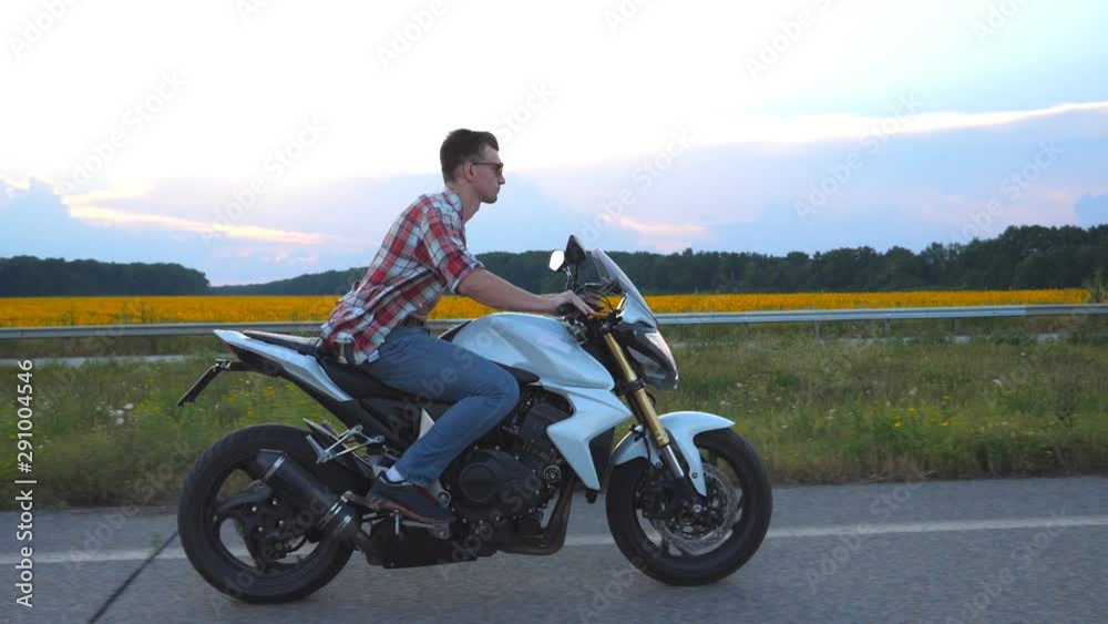 Young man riding on a modern sport motorbike. Handsome motorcyclist in a shirt and glasses driving his motorcycle on country road with sunflowers field at background. Slow motion Side view Close up Young man riding on a modern sport motorbike. Handsome motorcyclist in a shirt and glasses driving his motorcycle on country road with sunflowers field at background. Slow motion Side view Close up