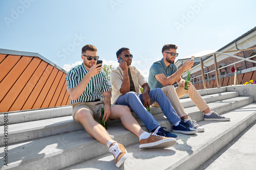 leisure, technology and people concept - happy male friends with smartphones drinking beer and talking on street in summer