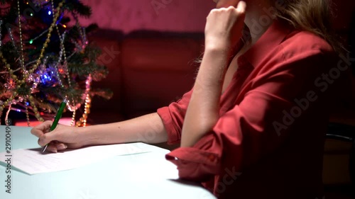 woman sits at a table near a decorated Christmas tree and writes a Christmas letter to Santa Claus photo