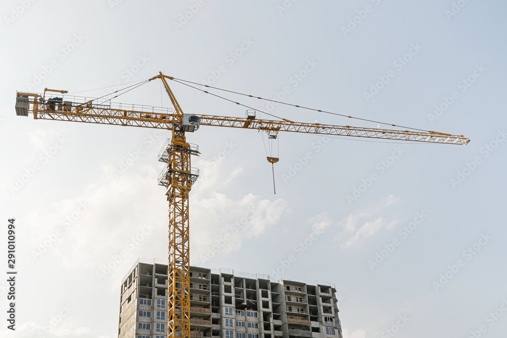 A yellow high-rise building crane against a blue sky builds multi-storey apartment buildings using modern technologies of metal, concrete and brick according to an architectural project