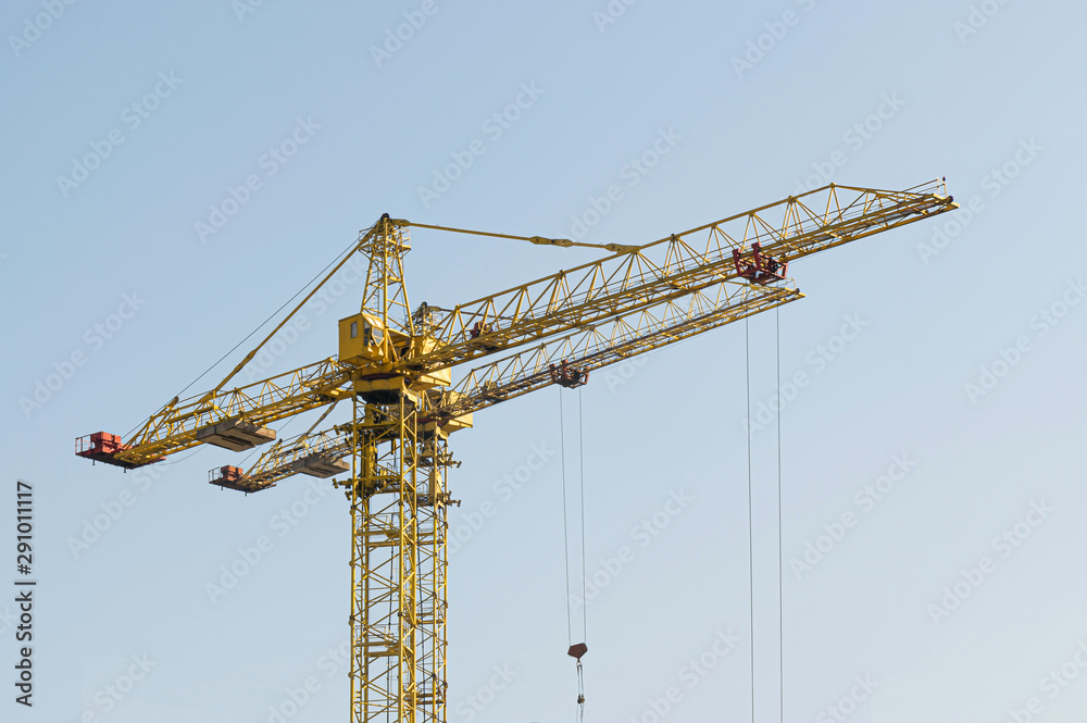 A yellow high-rise building crane against a blue sky builds multi-storey apartment buildings using modern technologies of metal, concrete and brick according to an architectural project A yellow high-rise building crane against a blue sky builds multi-storey apartment buildings using modern technologies of metal, concrete and brick according to an architectural project