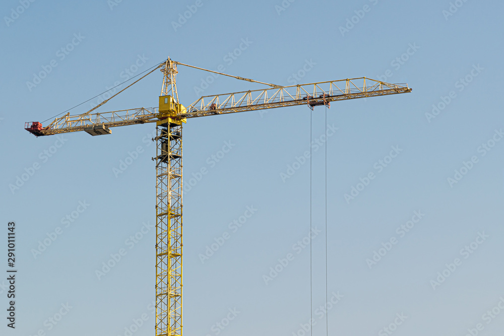 A yellow high-rise building crane against a blue sky builds multi-storey apartment buildings using modern technologies of metal, concrete and brick according to an architectural project A yellow high-rise building crane against a blue sky builds multi-storey apartment buildings using modern technologies of metal, concrete and brick according to an architectural project