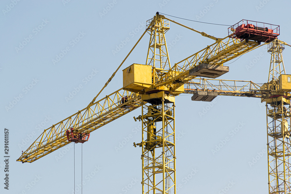 A yellow high-rise building crane against a blue sky builds multi-storey apartment buildings using modern technologies of metal, concrete and brick according to an architectural project A yellow high-rise building crane against a blue sky builds multi-storey apartment buildings using modern technologies of metal, concrete and brick according to an architectural project