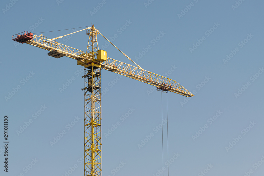 A yellow high-rise building crane against a blue sky builds multi-storey apartment buildings using modern technologies of metal, concrete and brick according to an architectural project A yellow high-rise building crane against a blue sky builds multi-storey apartment buildings using modern technologies of metal, concrete and brick according to an architectural project