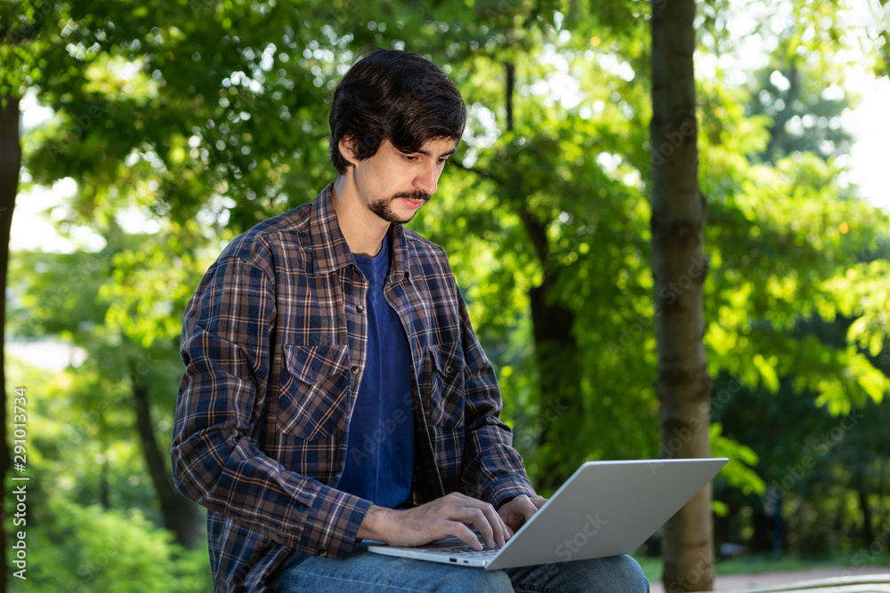 Young bearded brunet student with mustache sitting with a laptop in a park. Distance learning