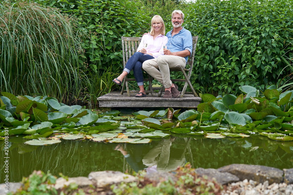 Portrait Of Mature Couple Celebrating With Champagne Sitting On Chairs On Wooden Jetty By Lake