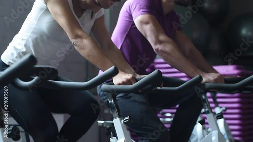 Attractive woman and man biking in the gym, exercising legs doing cardio workout cycling bikes. Couple in a spinning class wearing sportswear. Fitness healthy lifestyle concept