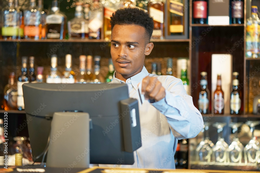 African bartender registration of a new order by a cash register. A restaurant worker registrating new order by cash-register. The barman pays the order with a credit card.