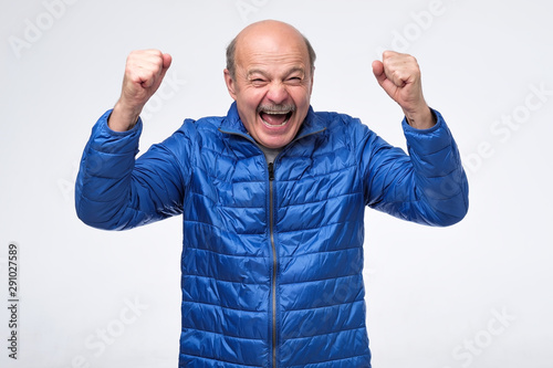 Lucky successful senior man in blue jacket exclaiming, clenching fists, having winning look after showing the best results. Studio shot photo