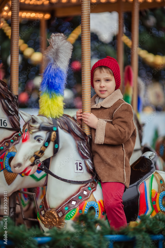 Cute smiling boy in a brown coat and a red hat rides on a merry-go-round.