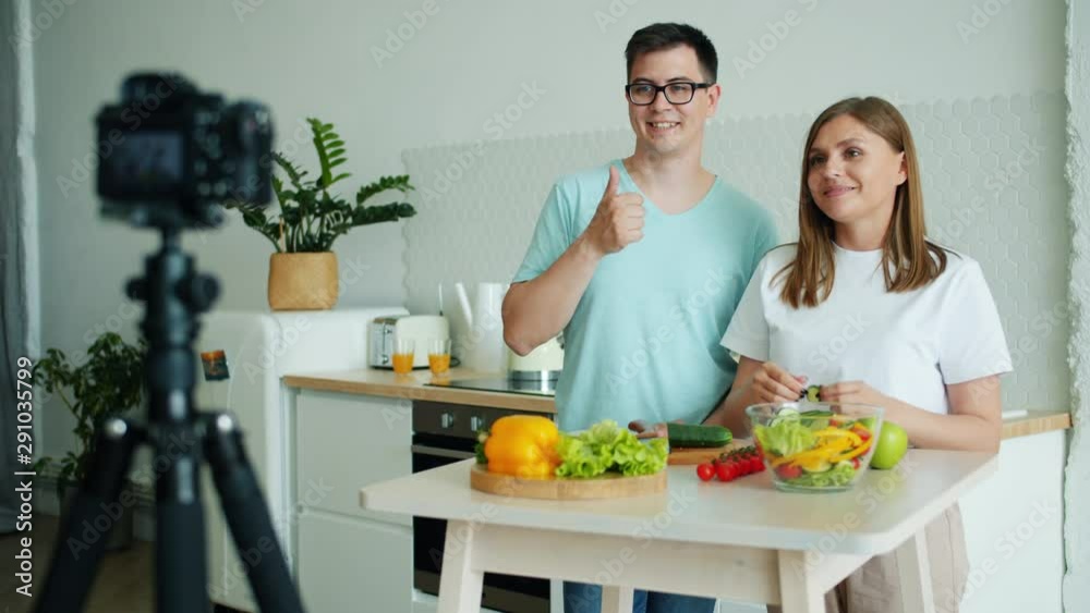 Couple of blogger young attractive man and woman are recording video in kitchen showing thumbs-up waving hand smiling using professional camera on tripod. Couple of blogger young attractive man and woman are recording video in kitchen showing thumbs-up waving hand smiling using professional camera on tripod.
