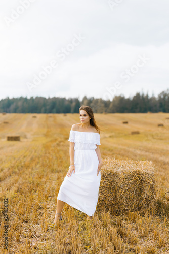 Beautiful girl in a long white dress and barefoot standing at setga of straw on a yellow field in summer