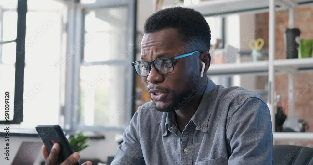 Happy African American man using smartphone having video chat enjoying online communication indoors. Man in glasses holding phone and have online video call in modern office. Happy African American man using smartphone having video chat enjoying online communication indoors. Man in glasses holding phone and have online video call in modern office.