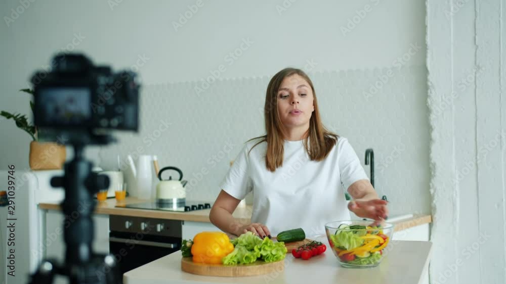Slow motion of pretty lady recording video about vegetables in kitchen at home cooking salad talking waving hand using camera. Lifestyle and vlogging concept. Slow motion of pretty lady recording video about vegetables in kitchen at home cooking salad talking waving hand using camera. Lifestyle and vlogging concept.
