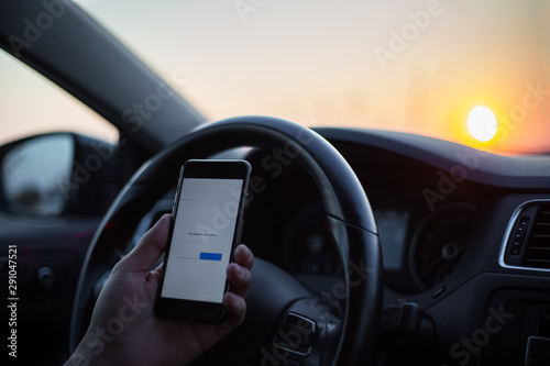 Close-up of male hand hold smartphone inside car against steering wheel at sunrise. No network connection.
