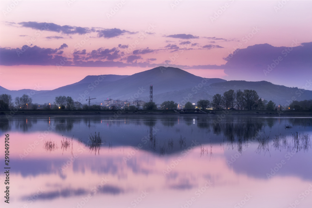 Distant mountain and city, blue hour lake mirror reflection in a calm water of vivid purple sky 